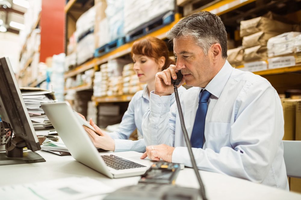 Manager working on laptop and talking on phone at desk in a large warehouse